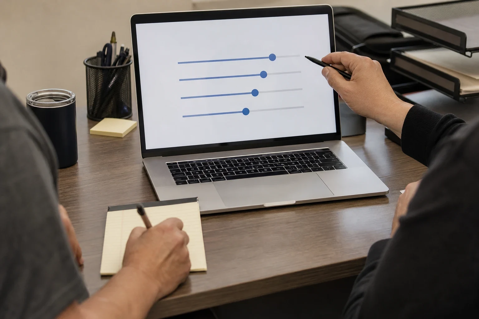 Two people at an office desk: one points a pen at adjustable sliders on a laptop screen while the other takes notes on a yellow legal pad