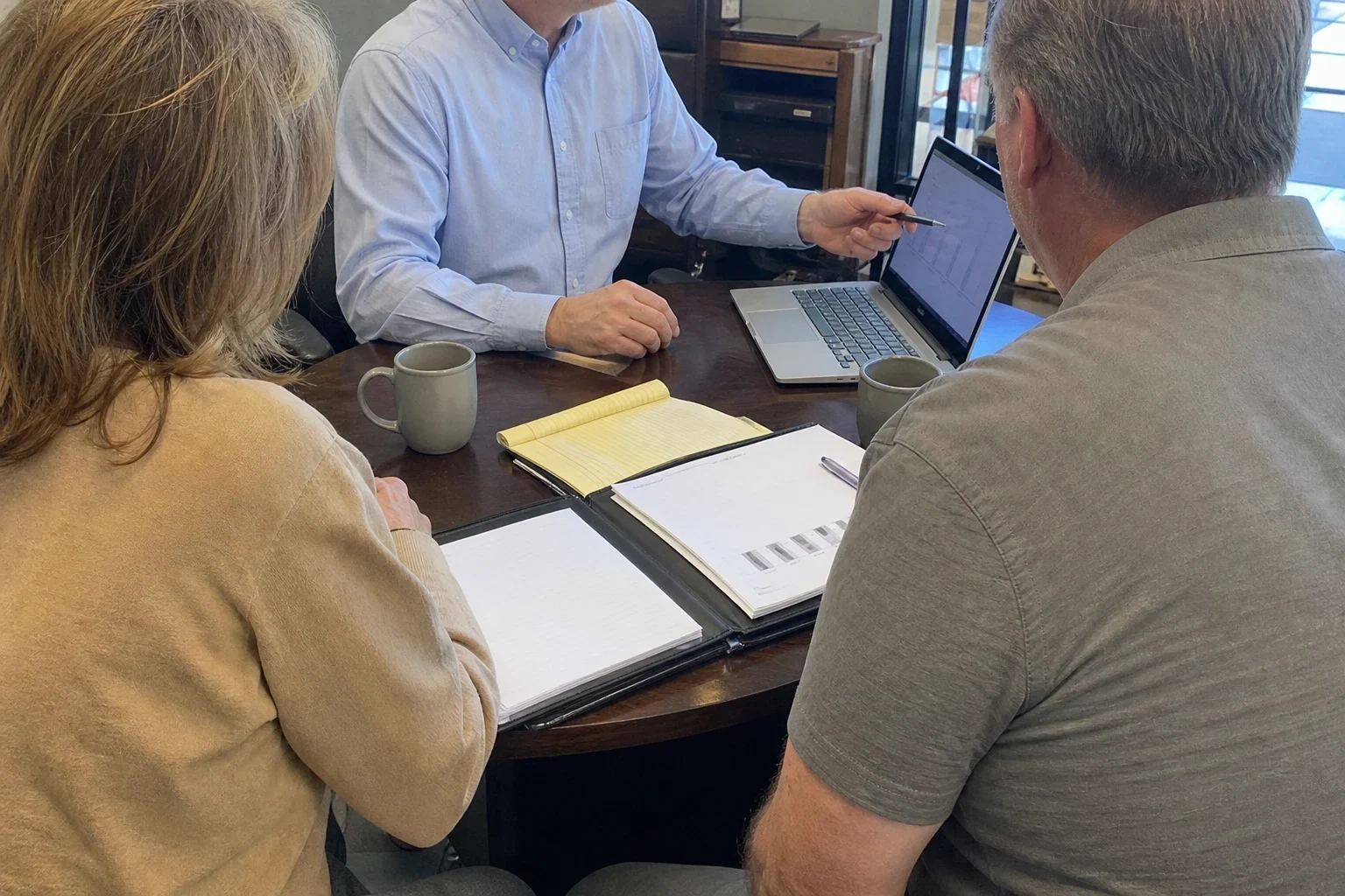 Agent in a light blue button-down shirt points a pen at a laptop while a couple reviews papers in a padfolio at a round wooden table in an office